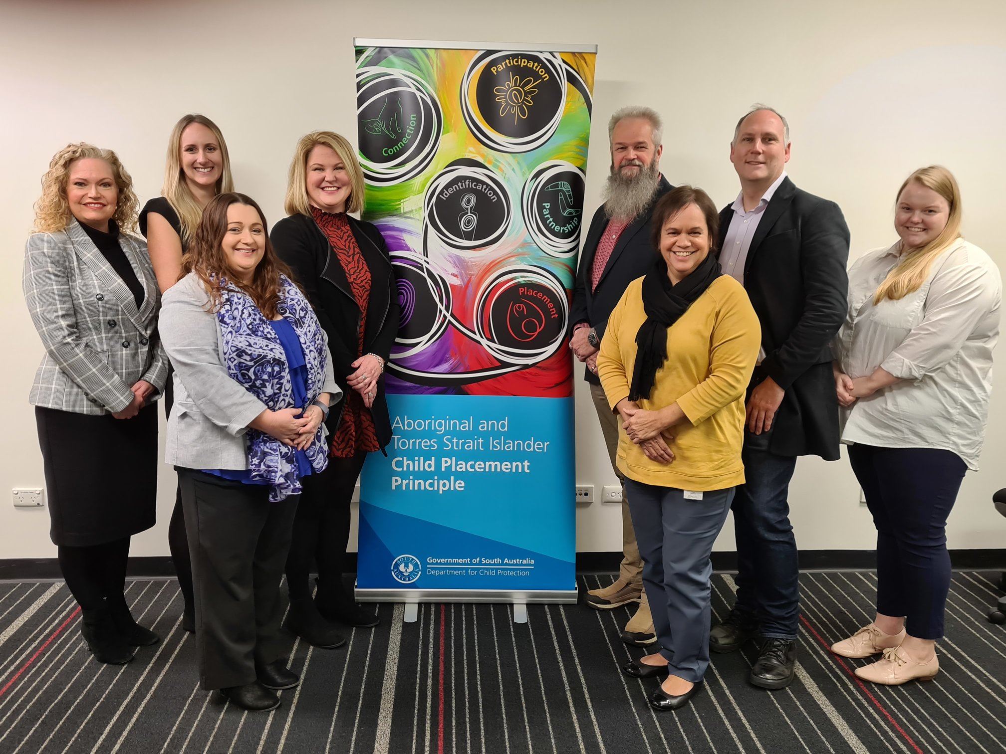DCP Procurement and Contracting, and Aboriginal Practice staff 8 Department for Child Protection staff stand near a pull up banner with a colourful representation of the Aboriginal Child Placement Principle.